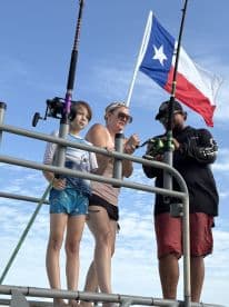 A family engaging in fishing on a boat, with a Texas flag waving in the background. The young boy and woman are focused on the fishing gear, showcasing a fun and interactive fishing experience.