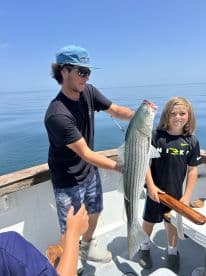 A young angler proudly displaying a large Striped Bass while fishing on a calm day at sea, accompanied by a friend.