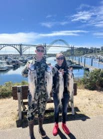 Two anglers proudly displaying their catch of large salmon at a scenic marina, with a bridge in the background.