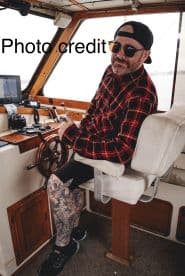 A man seated at the helm of a fishing boat, showcasing the interior design and controls, with a focus on his detailed tattoo work.
