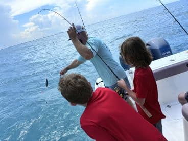 Two young anglers actively participating in a fishing trip, with one reeling in a fish while the other observes, aboard a boat in open water.