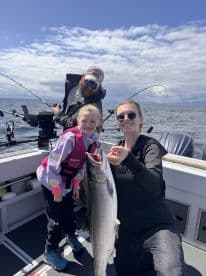 A young girl and her mother proudly display a large Salmon while fishing on a boat, enjoying a family outing in a scenic marine environment.