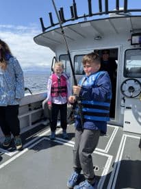 A young angler is actively reeling in a fish while wearing a life jacket on a fishing boat, surrounded by fellow young fishers.