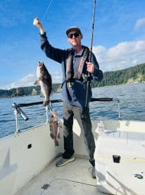 Angler proudly displaying a catch of two large rockfish while fishing from a boat in a scenic coastal environment.