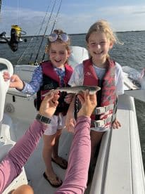 Two young girls excitedly display a small shark while fishing on a boat, showcasing a fun family fishing experience.