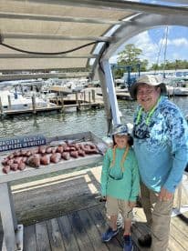 A young angler and an adult proudly pose beside a catch of Red Snapper displayed on a cleaning table at the marina.