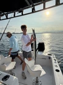 A young angler proudly displaying a Barracuda on a fishing boat during a sunny day on the water.