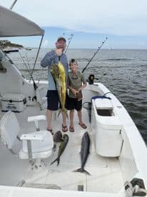 A proud young angler and an adult display their catch, including a large Mahi Mahi and other fish, aboard a fishing boat in a coastal setting.
