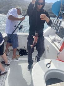 Angler reeling in a colorful rockfish while fishing on a charter boat in a scenic coastal environment.
