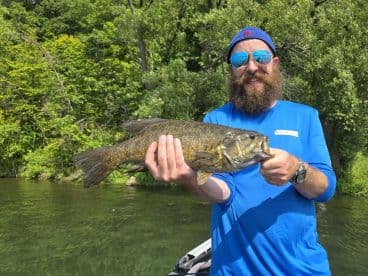Angler proudly displaying a large Smallmouth Bass while fishing in a scenic freshwater environment.