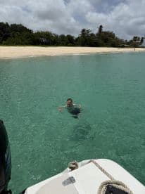 A young boy enjoying a swim in the clear waters near a sandy beach, with a boat visible in the foreground.
