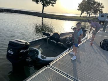 A young boy in a life jacket stands beside a sleek fishing boat at the dock, with a serene waterfront view in the background.