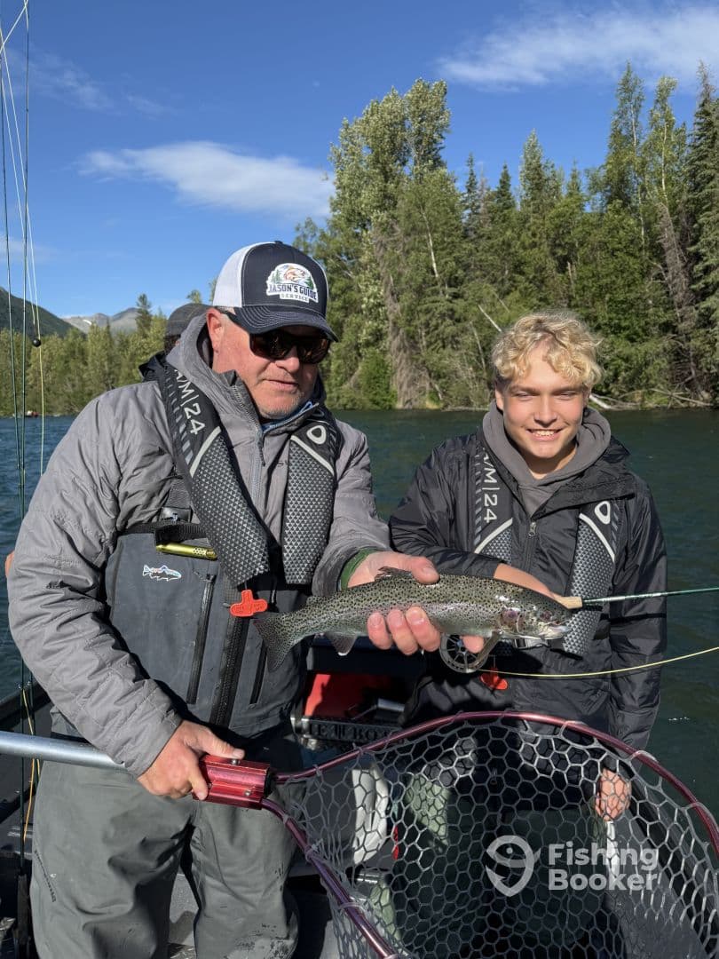 Two anglers proudly displaying a Rainbow Trout while fishing on a scenic river, showcasing a successful catch during their trip.
