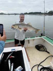 Angler proudly displaying a large Striped Bass while fishing from a boat, showcasing a successful catch in a scenic waterway.