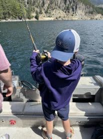 A young angler is actively reeling in a fish while fishing on a boat in a scenic lake surrounded by mountains.