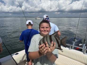 A young angler proudly displaying a striped fish while fishing on a boat, surrounded by fellow anglers on a sunny day.