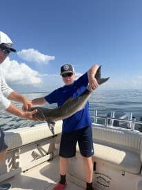 A young angler proudly displaying a large shark on a fishing charter, surrounded by calm waters and a clear blue sky.