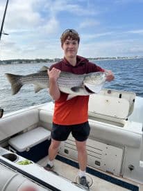 Young angler proudly holding a large Striped Bass aboard a boat, showcasing a successful fishing trip.