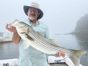An angler proudly displaying a large Striped Bass while fishing in a foggy coastal environment.