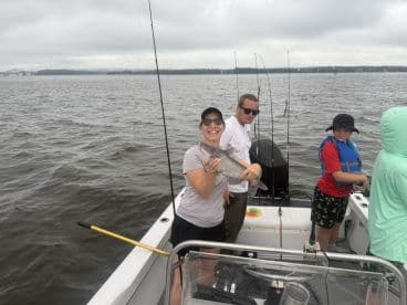 A group of anglers enjoying a fishing trip, with a woman proudly displaying a caught catfish while others assist on a boat in a river setting.
