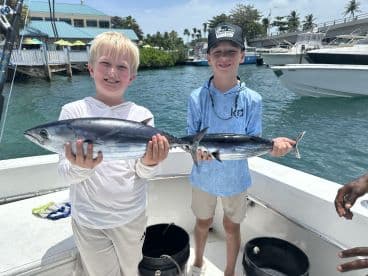 Two young anglers proudly displaying their catches of Bonito on a fishing charter in a tropical setting.