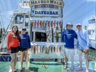 A family poses in front of their catch at the HarborWalk Marina, showcasing a variety of fish displayed on a board after a successful fishing trip.