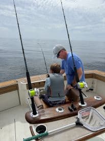 A young angler in a life jacket receives guidance from an adult while fishing on a boat, showcasing a calm day on the water.