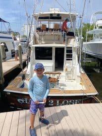 A young boy stands on the dock in front of the 'Game 2 Lose' fishing boat, showcasing the boat's exterior and inviting atmosphere for family fishing adventures.