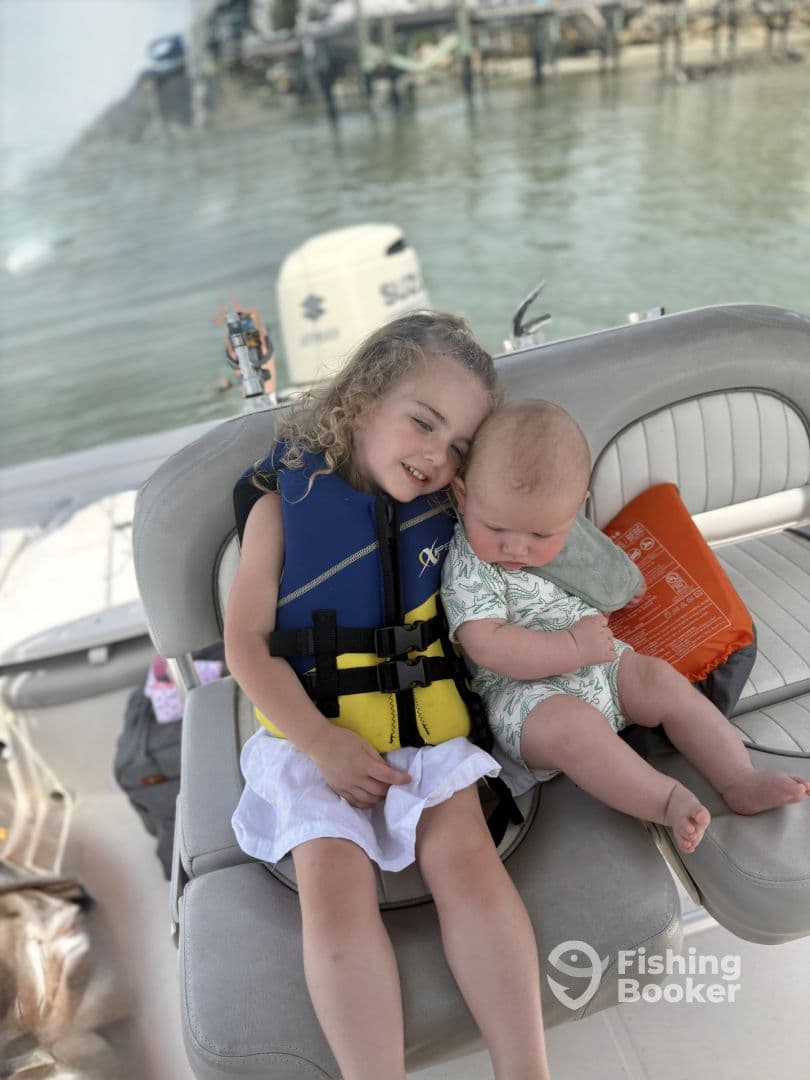 Two young children enjoying a day on the boat, seated comfortably in life jackets, with a scenic waterfront in the background.