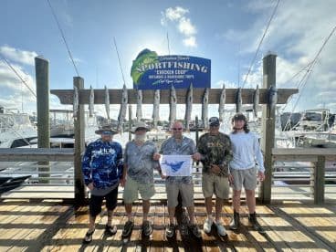 A group of five anglers proudly displaying their catch of Barracuda on a catch board at a marina, showcasing a successful offshore fishing trip.