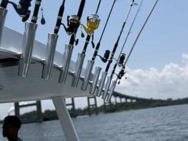 A close-up view of fishing rod holders on a boat, with a young angler in the background, showcasing the fishing setup against a scenic waterfront and bridge.