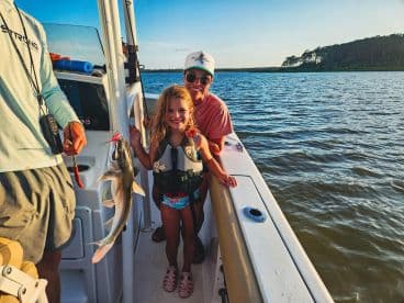 A young girl proudly displays her catch, a small fish, while enjoying a family fishing trip on a boat in a scenic waterway.