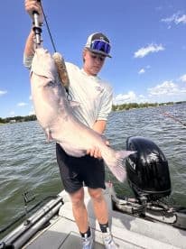 Young angler displaying a large Catfish while fishing on a lake, showcasing a successful catch during a sunny day.
