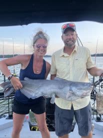 Two anglers proudly displaying a large Blue Catfish while fishing on a lake, showcasing a successful catch during their trip.