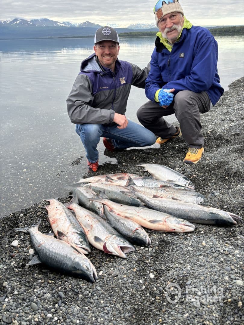 Two anglers proudly displaying a catch of multiple Salmon on a rocky shore, with a scenic backdrop of mountains and water.