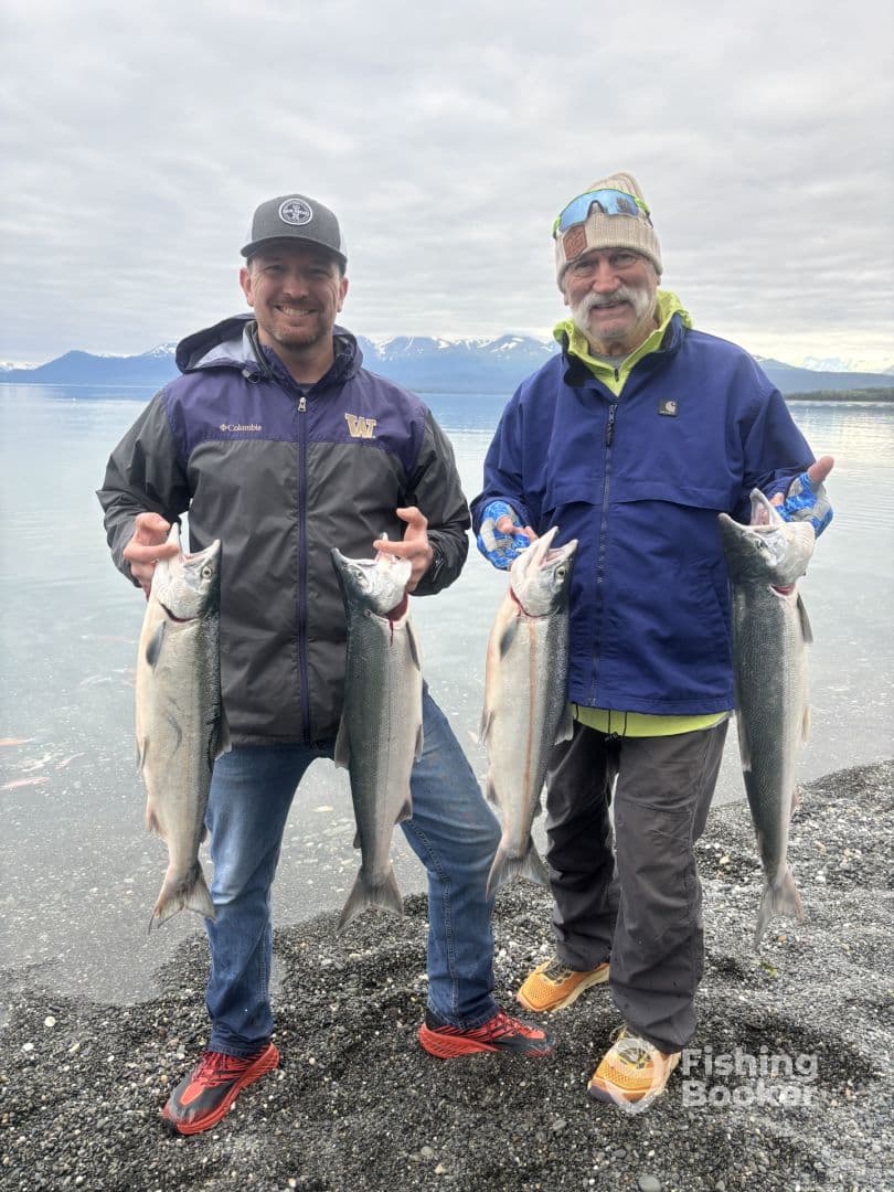 Two anglers proudly displaying their catch of Silver Salmon on a rocky beach with a scenic mountain backdrop.