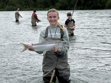 A young angler proudly displaying a Silver Salmon while wading in a river, surrounded by fellow fishermen enjoying a day of fishing.
