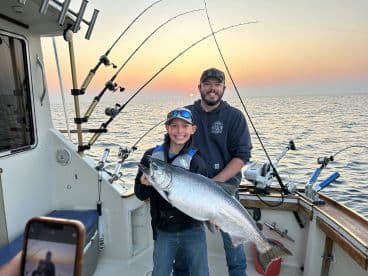 A young angler proudly displaying a large Salmon while fishing on a boat during sunset.