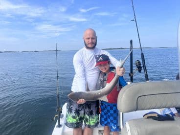 A proud father and son duo display a caught shark while fishing on a boat, showcasing a fun family fishing experience.