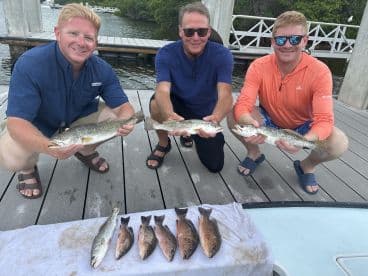 Three anglers proudly displaying their catch of Speckled Trout on a dock after a successful fishing trip.