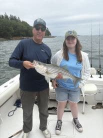 A proud angler and a young girl displaying a large Striped Bass while fishing on a boat in a scenic coastal environment.