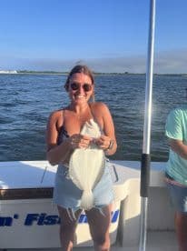 A smiling angler proudly displays a Flounder while fishing on a boat, enjoying a sunny day on the water.