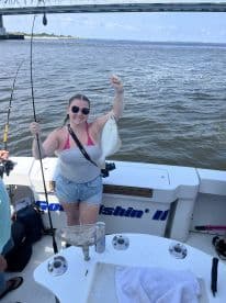 A smiling angler proudly displays a caught Flounder while fishing on a boat, with a scenic backdrop of a bridge and water.