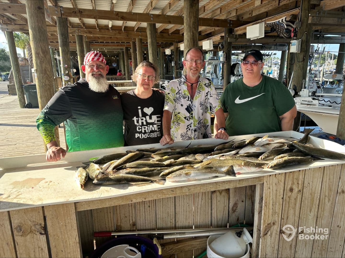 A group of four anglers proudly displaying their catch of Speckled Trout on a catch board at a marina, showcasing a successful fishing trip.