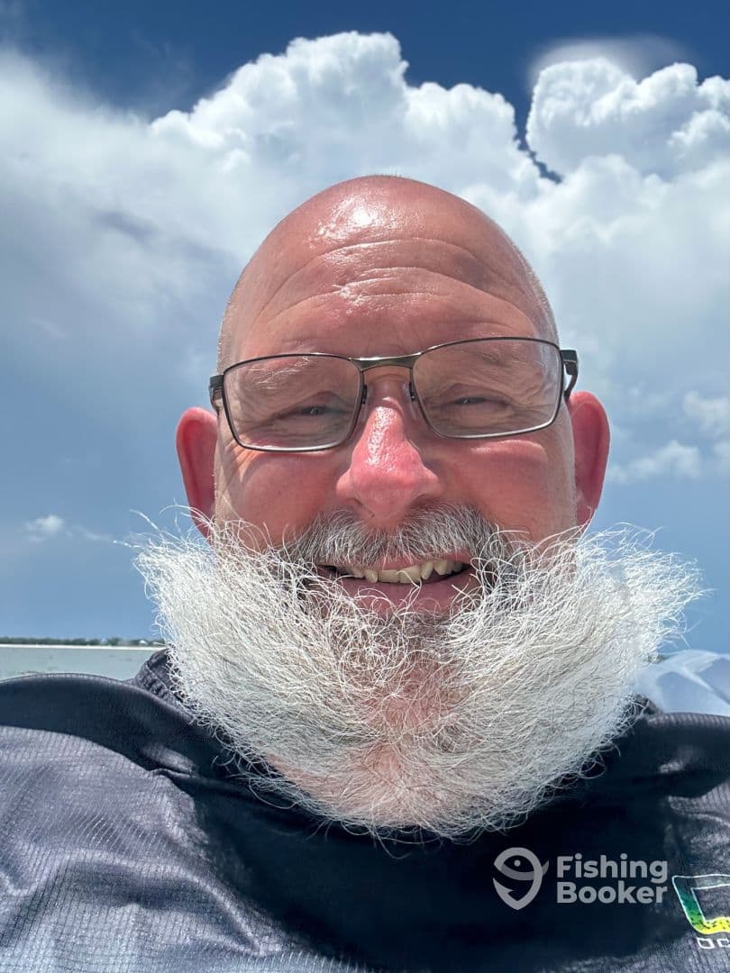 A smiling individual with a distinctive white beard, enjoying a sunny day on a boat with a backdrop of fluffy clouds.