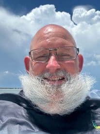 A smiling individual with a distinctive white beard, enjoying a sunny day on a boat with a backdrop of fluffy clouds.