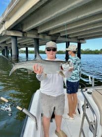 Young angler proudly displaying a large Tarpon while fishing under a bridge, with another child in the background.