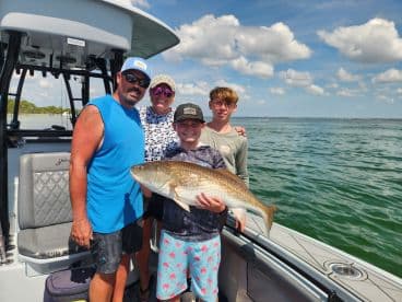 A family proudly displays a large Redfish on a boat during a sunny day on the water.
