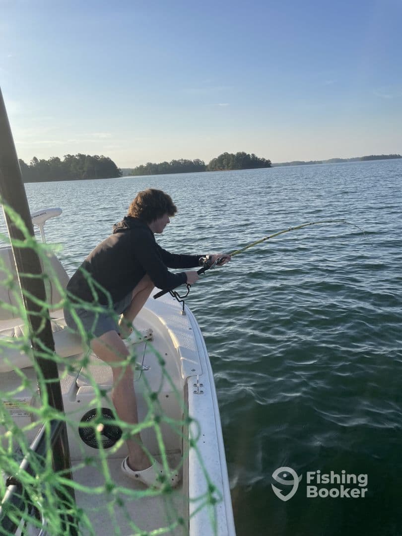 A young angler is actively reeling in a fish from the side of a boat on a serene lake, surrounded by lush greenery.