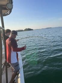 Two anglers engaged in fishing action on a boat, with one preparing to net a catch in a scenic lake setting.